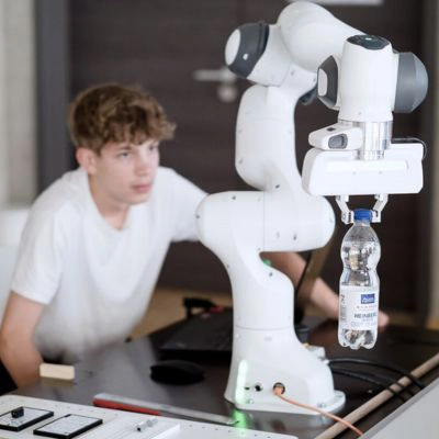 Junger Auszubildender beobachtet kollaborativen Roboterarm, der eine Wasserflasche greift – praxisorientierte Lernumgebung.