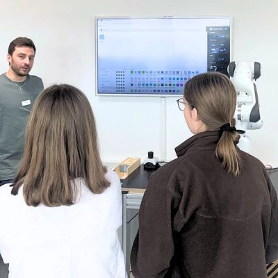 Zwei Auszubildende beobachten eine Bildschirmpräsentation zur Robotersteuerung in einem Schulungsraum.