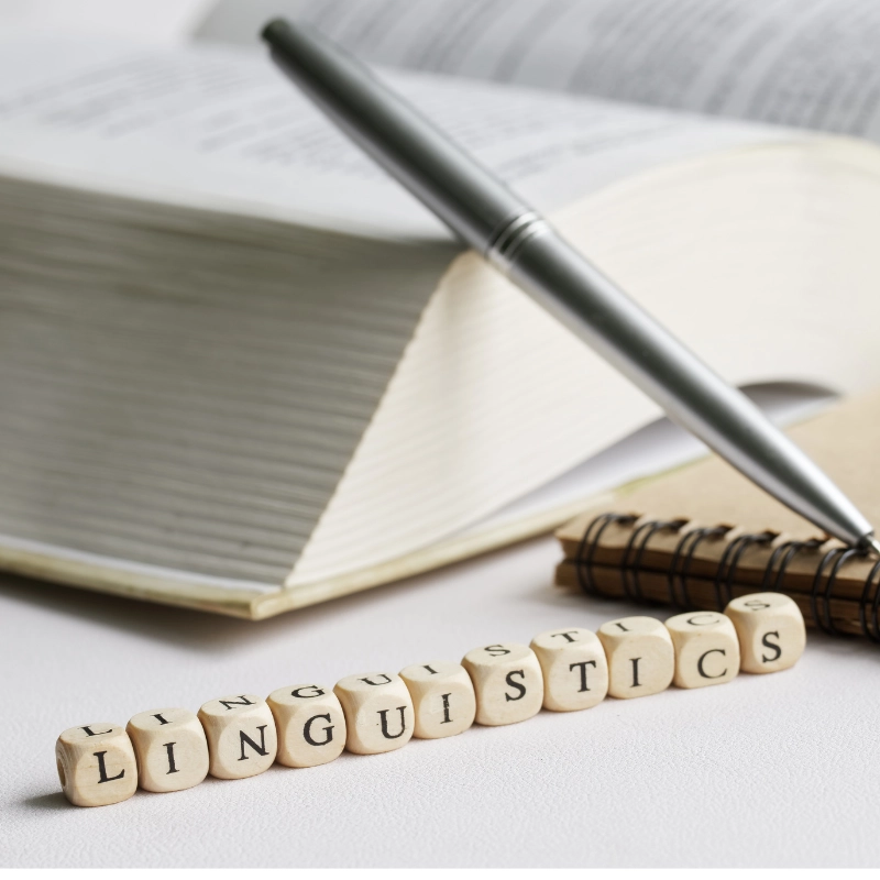 Nahaufnahme eines Wörterbuchs mit Notizbuch, Stift und Holzbuchstaben, die das Wort „Linguistics“ bilden – Symbol für Sprachwissenschaft und Rhetorik.