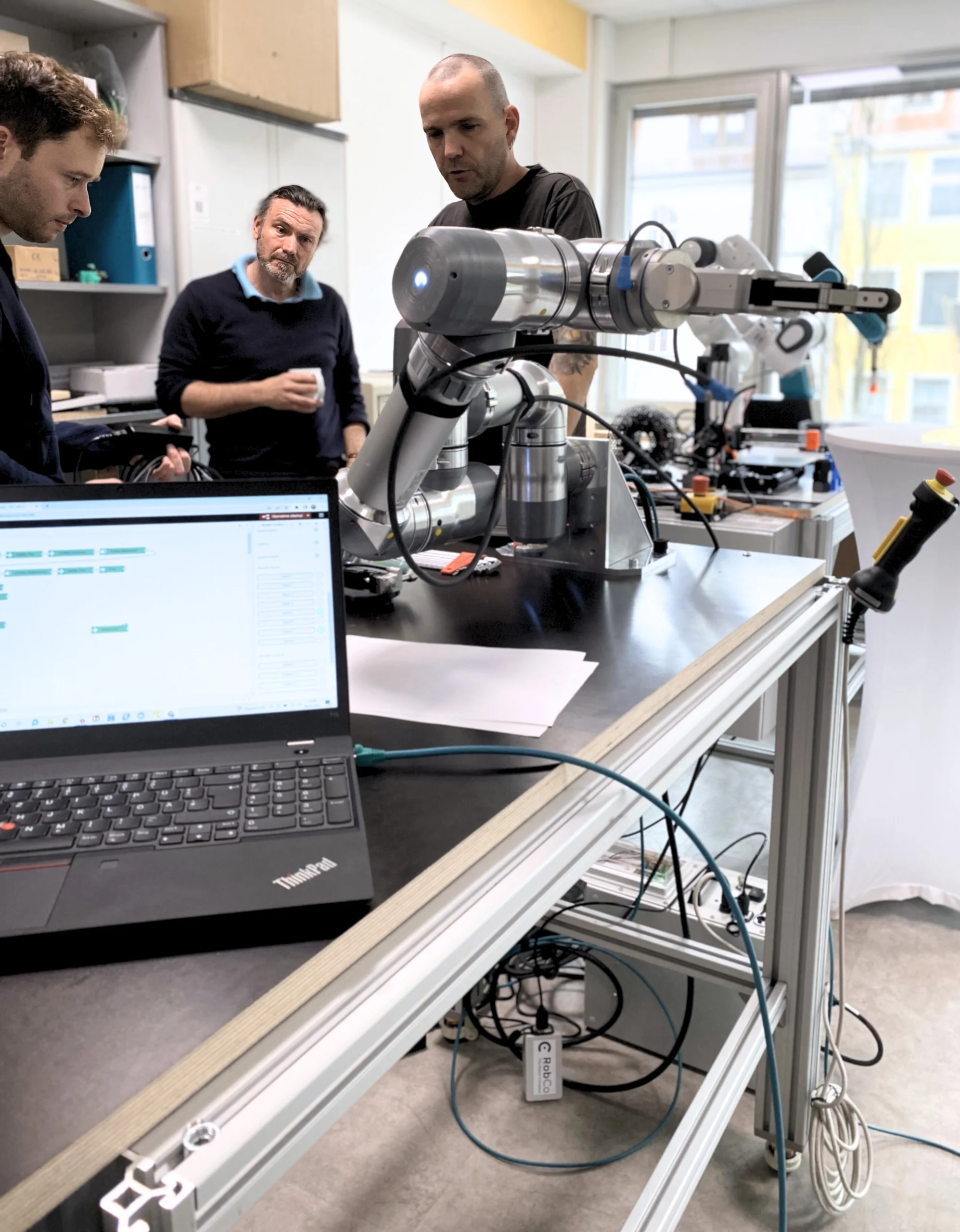 Drei Männer arbeiten gemeinsam mit einem kollaborativen Roboterarm an einem Programmierarbeitsplatz – Weiterbildung in der Industrie.