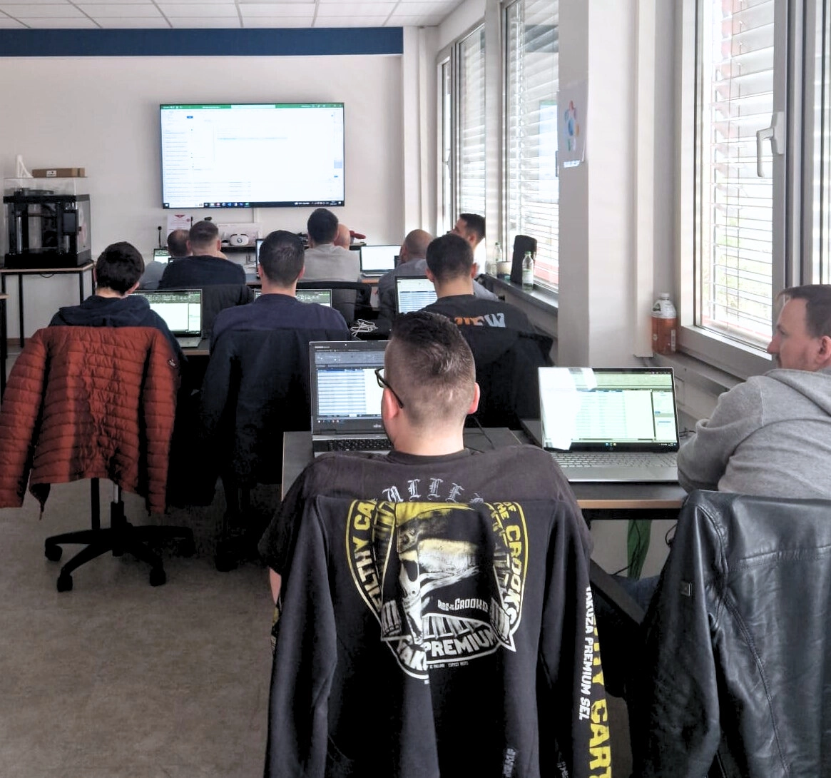 Teilnehmer sitzen mit Laptops in einem modernen Schulungsraum und verfolgen digitale Inhalte auf einem großen Bildschirm.